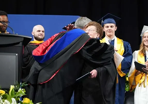Sister Mary Ann Stewart hugging Cathedral High School President, Dr. Robert Bridges, after receiving her honorary diploma as Cathedral teachers (back, from left) Matt Moorman and Danny Goggans along with Cathedral 2026 graduates James Grimme and Lauren Robertson look on. (Photo courtesy of Inter-State Studio).